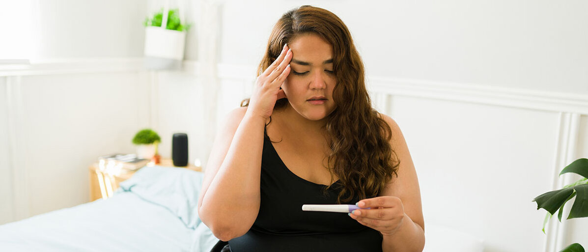 Eine mehrgewichtige Frau sitzt auf der Bettkante und hält einen Schwangerschaftstest. Sie schaut entsetzt.